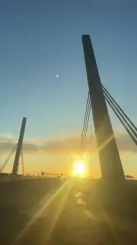 The video shows a low-angle view of a bridge at sunset. Two tall, concrete towers stand on either side of the road, supporting the bridge with thick cables. The sun shines directly ahead, creating a bright glare and long shadows on the road. Several cars are visible in the distance. The sky is blue with wisps of clouds near the horizon. The camera appears to be moving forward, suggesting the video was taken from a vehicle driving across the bridge. Music plays in the background.