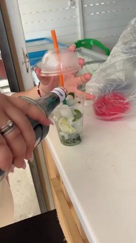 A close-up shot of a person preparing a drink on a white countertop. The person is wearing a white top and has light pink manicured nails and a silver ring with a clear stone on their left ring finger. They are holding a clear plastic cup with a dome lid and an orange straw in their hands. The cup contains ice cubes, green mint leaves, and slices of lime. The person is holding a light blue bottle with a red cap in their right hand, appearing to pour liquid into the cup. Another clear plastic bag with a red object inside is visible on the counter to the right. The background shows part of a light-colored wall or cabinet and some shelves with items.