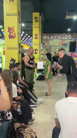 A muscular man in a black tank top and green pants stands near a yellow pillar decorated with cat images. He holds a white bag in his left hand and appears to be interacting with a woman in a green outfit. The woman is holding a phone and a microphone. Several other people are visible in the background, some seated and others standing. The setting appears to be an indoor public space, possibly an entertainment center, with colorful decorations on the ceiling and a "PLAY KIDS" sign in the background. The man is smiling, and the overall atmosphere seems lighthearted.