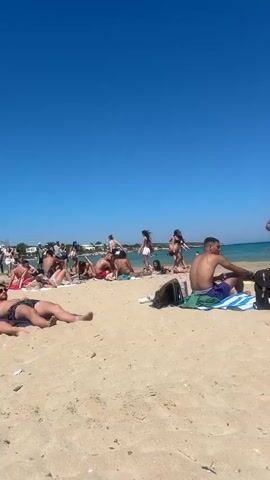 The video shows a crowded beach scene on a sunny day with a clear blue sky. In the foreground, two people are visible on the sand, one sitting with a backpack beside them, and another standing and adjusting their clothing. In the background, a group of men are gathered, and further down the beach, more people are seen sunbathing and interacting. The water appears calm and is a light blue color. The environment is sandy with some scattered beach accessories like towels and flip-flops.