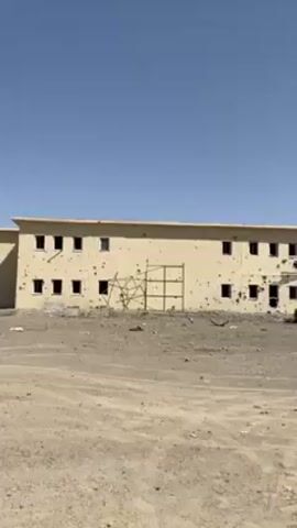 The video shows a two-story building with many bullet holes and other damage on its facade. There is a damaged car parked in front of the building, amidst rubble and debris. A makeshift structure made of scaffolding stands near the damaged entrance. The environment is a barren, desert-like landscape under a clear blue sky. The camera slowly pans to the right, revealing the extent of the damage to the building and the surrounding area.
