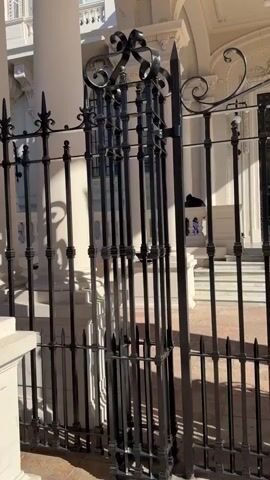 The camera pans upwards to reveal a tall, white, ornate building behind a black iron gate. A person in dark clothing stands on the porch of the building. The building has columns and arched entrances, and ornate ceiling light fixtures can be seen. The sky is bright and the sun casts shadows from the gate onto the building.