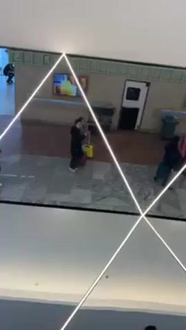 From a low angle looking upwards, the video shows a ceiling with crossing white neon lights that reflect a scene below. The reflection shows a hallway with a marble-like floor and a wall with two televisions displaying moving images.  Several people are seen walking in different directions, some pulling luggage.  A service cart, a trash can, and a closed door are also visible along the wall.  A dining area with tables and people is partially visible at the far right of the reflection.  Two people are walking together, one pulling a yellow suitcase and waving, as they approach the foreground of the reflected scene.