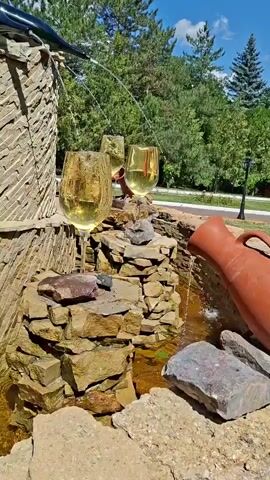 A close-up shot captures a decorative fountain crafted from stacked stones and a terracotta jug. Three large wine glasses filled with a golden liquid, likely wine, are strategically placed on the stones, with water from the fountain gently pouring into them and overflowing. The background reveals a lush green environment with trees and a blue sky with scattered clouds, suggesting an outdoor setting.