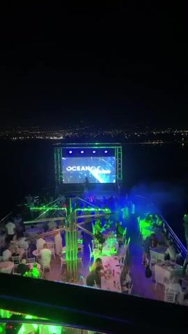 A night time aerial view of a party on a boat. A large LED screen with changing graphics is positioned at the front of the boat, with a DJ standing in front of it. The screen displays "OCEAN VIBRATIONS". Green and blue lights illuminate the deck, where numerous people are seated at tables and standing. Smoke drifts across the scene. The water surrounds the boat, and a distant city skyline is visible.