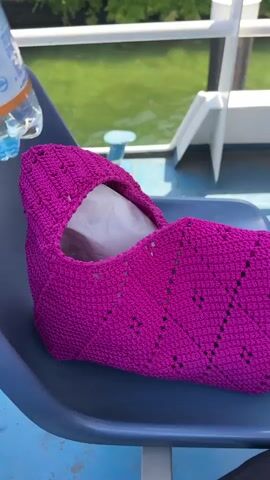 A person's hand, adorned with a pink nail polish and a ring, is placing a plastic bottle of RECOARO acqua minerale naturale into a crocheted bag. The bag is a vibrant fuchsia color with a textured, diamond-like pattern. It sits on a dark blue chair, likely on a boat or ferry, given the glimpse of green water and white structures in the background. The sun appears to be shining, casting a bright light on the scene. The overall mood seems relaxed and casual, possibly indicating a leisurely day trip or travel. The text on the bottle is clearly visible, identifying the brand and type of water. The setting suggests an outdoor environment, possibly during daylight hours.
