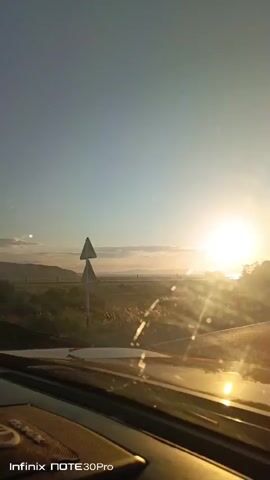 The video captures a view from inside a vehicle, looking out through the windshield. The sun is setting or rising, casting a bright glare and lens flares across the scene. The sky is a clear blue, transitioning to warmer hues near the horizon. In the distance, there are rolling hills or mountains partially obscured by mist or low clouds. The foreground shows the reflective surface of the car's dashboard or hood, with sunlight glinting off it. The surrounding environment appears to be a rural or semi-rural landscape with scattered vegetation and possibly a road. The Infinix NOTE 30Pro watermark is visible in the bottom left corner. There are no people visible in the frame. The video is shot from a moving vehicle, indicated by the slight blur and changing perspective of the scenery.