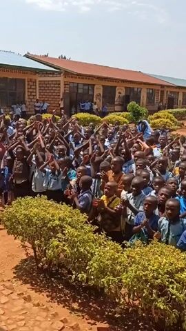 In an outdoor scene under a bright sky, a large group of elementary school children, mostly wearing blue and white uniforms, stand closely together, clapping their hands and raising their arms in what appears to be a celebratory or enthusiastic display. In the foreground, green bushes partially obscure the view. Behind the children, a school building with a red-tiled roof is visible, and in the background, the Rwandan flag is seen waving from a flagpole. A woman with dark hair is standing among the children and also clapping. The ground is a reddish-brown color, suggesting a dirt or clay surface.