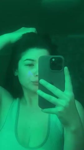 A young woman with dark hair, wearing a light-colored tank top, takes a selfie in a mirror. She has a blue mark on her cheek. The scene is illuminated by a green light, casting a shadow of her on the wall. Her right hand is raised and touching her hair. She is holding a dark-colored smartphone in her left hand, with the camera facing her.