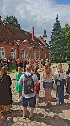 The video shows an outdoor performance with a large group of musicians playing instruments in front of a brick building. Several people are standing and watching the performance on a cobblestone street. A tall evergreen tree is on the right side of the frame, and a white church steeple is visible in the background. The sky is partly cloudy. The perspective is from behind the spectators looking towards the musicians.