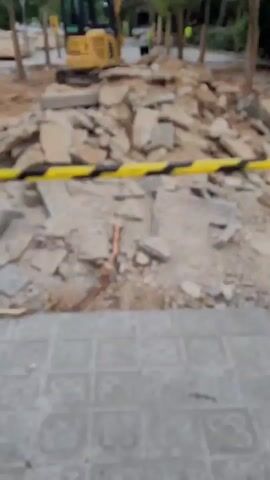 The video depicts an outdoor construction site, likely a pathway or public area under renovation. In the immediate foreground, the ground is covered with gray patterned concrete tiles, some intact, featuring a distinctive "Panot de Flor" (Barcelona flower) design. A yellow and black safety barrier stretches across the mid-ground, separating the viewer from the active work zone.

Behind this barrier, a substantial pile of construction debris is visible, composed of broken concrete slabs and rubble. Many of these broken pieces also display the "Panot de Flor" pattern, indicating the removal of old paving.

Further back in the scene, a yellow excavator, with "KOMATSU" clearly visible on its side, is positioned among more debris and trees. Two individuals, appearing to be construction workers in high-visibility vests, are present in the background near the excavator.

The scene suggests active demolition or clearing work. The time of day is daylight, and the weather appears dry. The yellow and black barrier includes a small pictogram, likely a safety warning. The prominent "Panot de Flor" tiles serve as a strong location cue, consistent with the context of Esplugues de Llobregat, Spain, indicating the removal of typical Catalan urban paving.