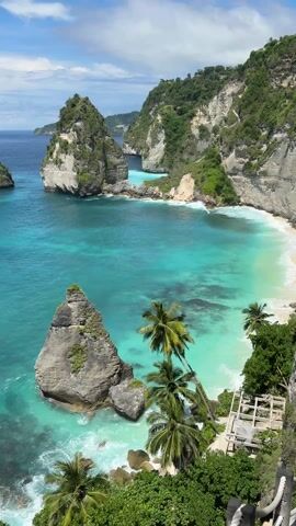 The video shows a scenic view of Diamond Beach on Nusa Penida Island in Indonesia.  The turquoise water of the ocean is visible, with waves crashing against the shore and rock formations. There are palm trees and lush greenery along the coast. A small wooden structure or platform can be seen near the edge of the viewpoint. The sky is partly cloudy with patches of blue.
