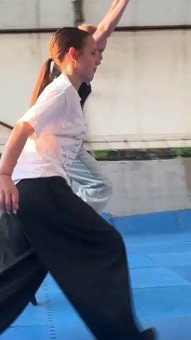 A group of young people are gathered on a blue mat-covered area, possibly a training space or dojo, under a white awning. In the foreground, a person is on their knees, facing away from the camera. To their left, another person is on the ground. In the center, a person in a white shirt and black pants is performing a martial arts move, possibly a cartwheel or a roll, with their body in mid-air. Further back, a group of individuals are watching the action. The environment appears to be outdoors, with trees and a building visible in the background. The lighting suggests it is daytime.