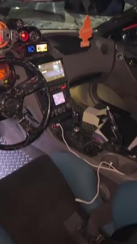 The video shows the interior of a modified car. The dashboard is visible, with a digital display, gauges, and a touchscreen infotainment system. The steering wheel is aftermarket, with a carbon fiber finish. The seats are racing style, with blue and black upholstery. The floor is covered with a diamond plate material. In the second part of the video, the hood of the car is open, revealing the engine bay. The engine appears to be modified, with a turbocharger and custom intercooler. The car is red with white and black accents. There are other cars and people visible in the background, suggesting the video was taken at a car show or event.