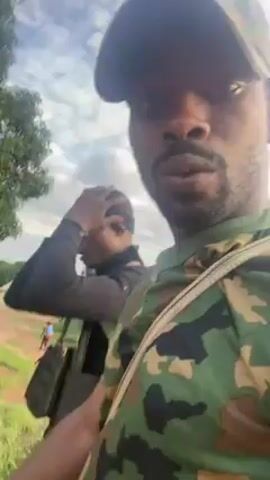Two men are outdoors. The man recording the video is wearing a camouflage shirt and a baseball cap. The other man is wearing a black long-sleeved shirt, a black beanie, and a tactical vest. He adjusts his beanie and smiles at the camera. Both men have equipment strapped to their shoulders. They appear to be standing on a grassy patch near a road. Trees are visible in the background.