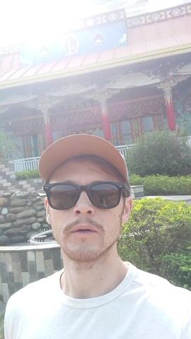 The video shows a selfie of a man in front of a temple. The man has a fair complexion, a light mustache and goatee, and is wearing sunglasses and a tan baseball cap. He’s dressed in a white t-shirt. The temple has a reddish-orange roof with a dome and what appears to be decorative carvings and paintings on its exterior walls. The temple also features red columns and a white railing. Behind the man and temple is a bright, sunlit sky. The surroundings of the temple include some greenery, a rock structure that appears to be part of a small fountain, and a few stone steps. The man points to the temple in the beginning of the video, and then holds the camera still facing the camera. He also appears to be talking to the camera, as his mouth is open.