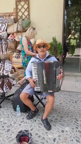 A man sits on a folding chair in front of a shop and plays an accordion. He is wearing a light blue button-down shirt, grey shorts, a straw hat, and sunglasses. The accordion is black and red. To his left is a black suitcase and a clear plastic water bottle. A basket sits on the ground in front of him, with a stuffed animal inside. In the background, there is a display of woven bags and baskets. A woman in a colorful dress walks past behind him. In the reflection of a shop window, another woman in a green dress can be seen. The ground is paved with cobblestones.