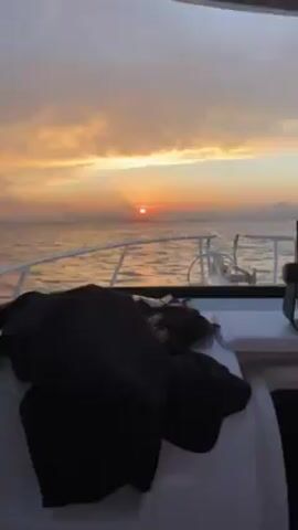 The video shows a sunrise over the ocean, viewed from the deck of a boat. The sky is a mix of orange, yellow, and gray clouds, with the sun peeking through the horizon. The ocean is calm with small waves, and the boat's white railing is visible in the foreground. A dark pile of clothing or gear sits on the deck.