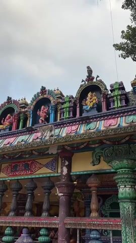 The video shows the ornate facade of a colorful Hindu temple against a cloudy sky. Several monkeys are visible on the roof of the temple. One monkey is walking along the ridge, while another is perched higher up on a decorative element. The temple's architecture is intricate, with multiple tiers of colorful decorations, statues of deities, and decorative pillars. The lighting suggests it is daytime, possibly overcast. The scene is viewed from a slightly low angle, looking up at the temple.