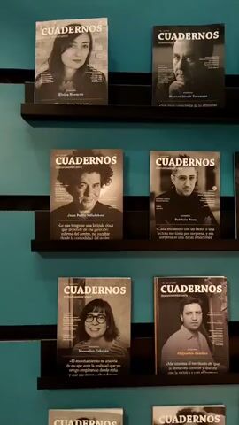 A video captures a wall painted in a solid teal color, adorned with multiple black shelves. Each shelf displays several magazines, all titled "CUADERNOS." Each magazine cover features a black and white portrait photograph of a person, typically a headshot or bust shot, with text below identifying the individual and a quote. The camera slowly pans upwards, revealing three rows of magazines on separate shelves, then reverses direction and pans downwards, showing the same magazines and an additional row at the bottom. The magazines are uniformly displayed, positioned upright with their covers facing forward.
