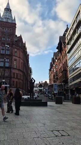 The video shows a street scene with several people walking and some architectural buildings. On the left side of the frame, there is a tall, red brick building with a distinctive tower and a camera mounted on it. In the center of the street, there is a statue of a person. On the right side, there are more buildings, one of which has the word "Cubo" visible on its facade. A double-decker bus is also present. Several people are walking in different directions. One man in a patterned shirt and shorts is walking towards the camera. Another man in a green polo shirt and blue shorts is walking away from the camera. A woman in a black top and jeans is also visible. The sky is cloudy with patches of blue. The ground is paved with rectangular stones.