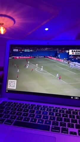 A person's hands are visible in the foreground, holding a laptop. The laptop is open, displaying a soccer game. The screen shows players in white and red uniforms on a green field. The score on the screen indicates ESP 1 - RSA 0. The game clock reads 16:31. The laptop is on a dark surface, possibly a lap or table. The background is dimly lit with purple and blue lighting, casting shadows on the ceiling. A light fixture is visible in the upper left corner. The atmosphere appears to be casual, suggesting the person is watching the game from home.
