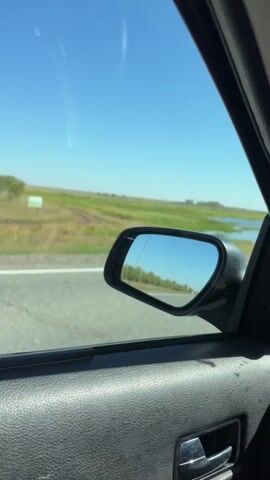 The video shows a view from inside a car, looking out the side window. The side mirror is visible, reflecting a road with trees and a blue sky. Outside the car, there is a grassy field with some water and a road. The sky is clear and blue. The car appears to be in motion.