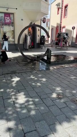 A video captures a street scene with a partial circular sculpture or fountain in the foreground. Paved with cobblestones, the ground has a mosaic of sunlight and shadow. To the left, a storefront displays clothing and accessories with a sign indicating "125 Jahre." A woman in white pants and a black top stands by the store window. To the right, racks of clothing are displayed outside a shop. A man walks past the sculpture, carrying a bag over his shoulder. Further right, a fountain with water splashing is visible. The background shows buildings with awnings and windows.