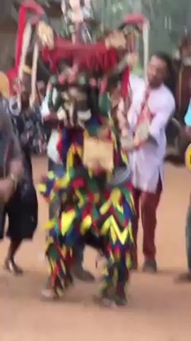 The video shows a person dressed in a colorful, elaborate costume and mask, apparently participating in a cultural performance. The costume is composed of overlapping shapes in primary colors, with a detailed mask featuring horns and tusks. Behind the costumed person, there are other people, some casually dressed and some appearing to be part of the performance with musical instruments. The ground is reddish-brown, suggesting a dirt or clay surface. The general atmosphere suggests a festive event or celebration outdoors.