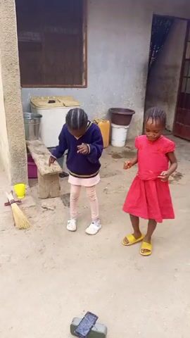 Two young girls are dancing outside near a building. The girl on the right wears a red dress and yellow sandals, and is smiling as she moves. The girl on the left is in a blue sweater, light pink leggings, and white shoes, looking down as she dances. A cell phone is on a speaker on the ground in front of them, presumably playing the music. The surroundings are a simple courtyard with various household items, including buckets, a broom, and a small bench. The building appears to be made of concrete or a similar material, and has a small, barred window.
