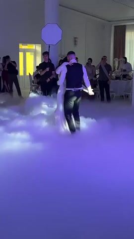 A wedding reception is underway. A couple, dressed in wedding attire, is dancing in the center of the room. They are surrounded by a thick cloud of fog, illuminated by purple lighting. Several other people are present, some are watching the couple, others are taking pictures. There are tables and chairs set up in the background, suggesting a banquet hall. A professional photographer with a large umbrella-shaped light modifier is positioned to the left of the dancing couple. Another person is crouched down to the left, also near the fog. The overall atmosphere appears festive and romantic.