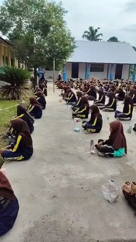 The video shows a group of young people, likely students, sitting on the ground in rows. They are dressed in uniform, consisting of brown hijabs and dark-colored uniforms with yellow accents. They appear to be gathered outdoors in a courtyard, possibly at a school, as a building with a white roof and blue trim is visible in the background. Some individuals have water bottles and other personal items placed near them. The scene suggests an organized gathering, perhaps for a lesson, assembly, or activity. The lighting indicates it is daytime, with an overcast sky.
