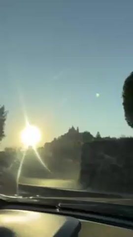 The video shows a drive. The camera looks out of a car's windshield as the sun shines brightly in the distance over what appears to be a building complex on a hill. A tree, a wall, and electrical poles pass as the car drives. A sign is seen on one of the poles.