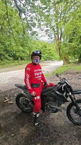 The video shows a person wearing a red riding suit and a helmet standing next to a black dirt bike in a wooded area. The person raises their arms in celebration and points to the river in the background. The environment includes green trees, a riverbed, and a dirt path. The lighting is bright, and the video is stable.