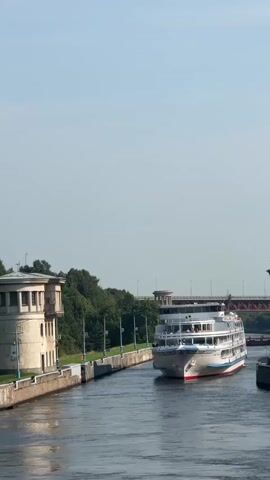 A white cruise ship with red and blue trim navigates a narrow waterway. The ship has multiple decks with passengers visible on them. On the left bank, a light-colored building with a tower is visible, and on the right bank, a similar tower structure stands. Trees line the left bank in the background. A red bridge with a complex structure is visible in the distance, spanning the waterway. The sky is clear and blue. The water is calm with gentle ripples. The lighting suggests it is daytime, likely a sunny day. The scene appears to be a lock or a canal system.