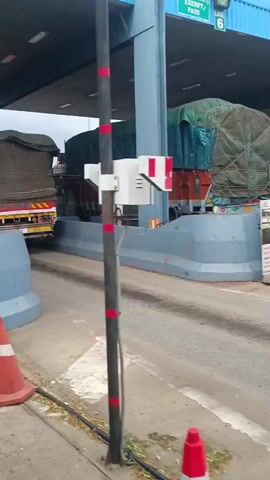 The video shows a view from a vehicle approaching a toll booth. Two large trucks, one in front of the other, are visible in the toll lanes. The trucks are covered with tarps, and their rear ends are facing the camera. The toll booth structure is overhead, with a pole and some equipment in the foreground. There is also an orange traffic cone on the ground. The overall environment appears to be an outdoor roadway.