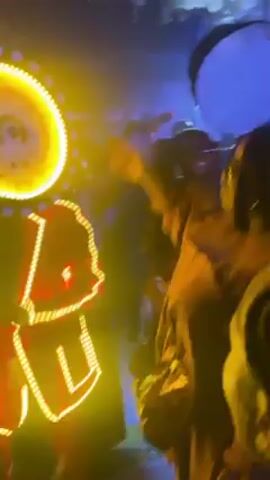 The video shows people in a crowded, dimly lit space, possibly a party or event.  Several individuals are visible, some wearing tan or orange clothing.  One person is adorned with a brightly lit LED costume, predominantly red and yellow.  People are moving around, interacting with each other, and recording the scene with their phones.  The lighting is varied, with bright spotlights and neon lights scattered throughout the room, creating a dynamic and lively atmosphere.
