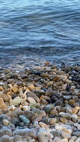 The video shows a close-up view of a pebble beach with gentle waves lapping at the shore. The water is a deep blue, with a lighter, more turquoise hue further out towards the horizon. In the distance, two people are visible in a kayak, paddling on the calm sea. The sky is a clear blue. The overall scene depicts a serene coastal environment on a sunny day.