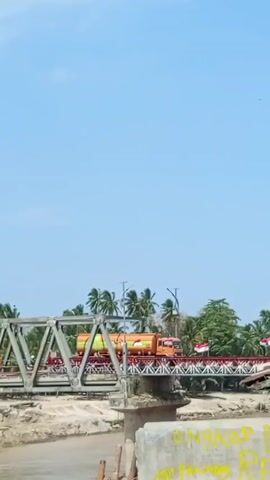 A brightly colored tanker truck crosses a silver steel-frame bridge over a shallow, muddy river on a clear day. Tropical trees and a small, dilapidated structure line the riverbank. The bridge is decorated with Indonesian flags. Graffiti is visible on the concrete support structures of the bridge.  The sky is clear and blue.