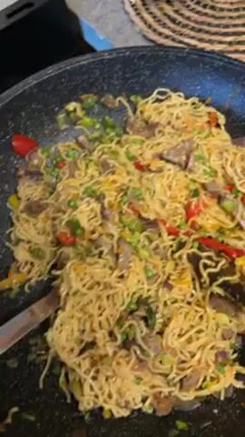 A close-up shot shows noodles being mixed in a large black pan using a spatula and a fork. The noodles are yellowish and mixed with pieces of meat, red bell pepper, yellow bell pepper, green onions, and peas. The pan sits on a stovetop next to a round woven placemat.