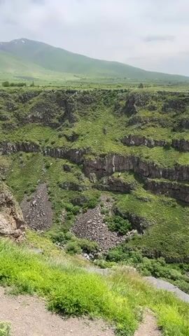 The video shows a canyon or gorge with stepped rock formations covered in grass and greenery. The opposite side of the canyon is a flat, grassy plain with a few small buildings visible. A large, green mountain is in the background under a cloudy sky. The viewpoint is from the edge of the canyon, with some grass and a path visible in the foreground. There is some wind.