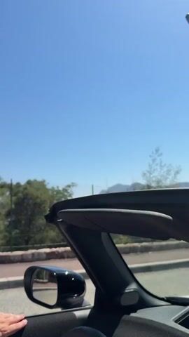 The video shows a view from the passenger seat of a convertible car. The roof is down, and the sky is clear blue. A hand is visible resting on the door frame. The car appears to be driving along a road next to a stone wall and a chain-link fence. Beyond the fence, there are trees, some vegetation, and in the distance, hills or mountains and possibly a body of water. The sun is shining brightly, creating a summery atmosphere.