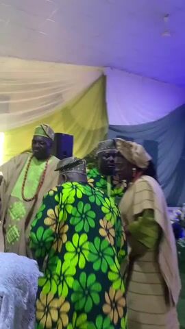 The video shows a small group of people, four visible, in an indoor setting possibly a banquet hall due to the draped ceiling decorations. The individuals are dressed in traditional African attire with vibrant green and gold patterns dominating. Two men wear matching caps and are adorned with beaded necklaces. A woman is also present in a similar head wrap and gown, smiling. Another woman in a green dress is visible in the foreground. The group appears to be in a celebratory mood. One of the men is holding what appears to be money in his hand. A black speaker is noticeable in the background, along with additional seating and people further in the backdrop. Overall, the scene seems festive, potentially part of a wedding or similar cultural celebration.