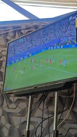 A flat-screen television displays a soccer match. The game is being broadcast on a channel called TVN. The scoreboard indicates that the game is in the 93rd minute. The players on the field are wearing red and white uniforms. The stadium is full of spectators wearing white. The television is mounted on a black stand with wires hanging from it. The background wall behind the television has a geometric pattern.