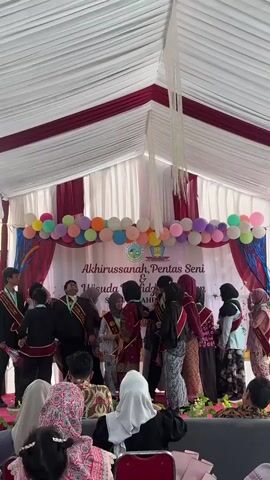 The video shows an outdoor event, possibly a graduation or awards ceremony, held under a white tent with maroon trim. A stage is set up with a backdrop featuring balloons and text that appears to read "Akhirussanah, Pentas Seni & Wisuda Tahfidz Qur'an". On the stage, a group of young people, likely students, are dressed in formal attire, some wearing sashes. Some are receiving awards or congratulations from adults. The audience is seated in front of the stage, with some individuals visible in the foreground wearing headscarves. Confetti is being released over the stage.