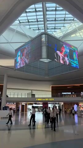 The video shows an indoor airport terminal. A large, multi-sided LED screen hangs from the ceiling, displaying colorful animated graphics with human figures. Several uniformed staff members in light-colored shirts and dark pants walk through the terminal. Other people, likely travelers, are also visible in the background. The terminal has a modern design with a high, glass-paneled ceiling and bright lighting. Stores and signage are visible along the walls.