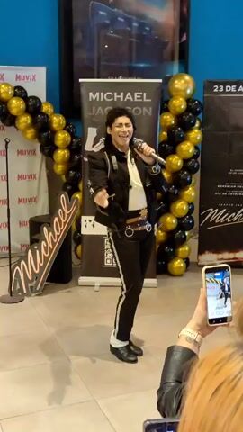A man dressed as Michael Jackson sings into a microphone. He is wearing a black jacket with silver detailing, black pants with white stripes, white socks, and black shoes. He has dark hair styled in a mullet. He is standing on a tiled floor in front of a banner that says "MICHAEL JACKSON". There are black and gold balloons arranged in an arch behind him. To his left, there is a sign with "Michael" written in red script. To his right, there is another banner with text that is partially visible. In the foreground, a person is holding up a smartphone to record the performance. The background appears to be an indoor space, possibly a cinema or event hall, with a movie poster visible on the wall.