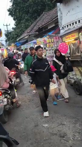 A busy street market is depicted with many people walking and riding motorcycles. The market stalls display various goods, including colorful balloons and other items. The people are dressed in casual clothing, and some are wearing headscarves. The atmosphere appears lively and crowded, suggesting a popular gathering place. The video captures the hustle and bustle of the market, with motorbikes navigating through the throngs of people.