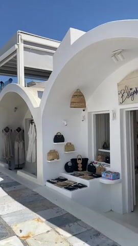 A short video taken on a sunny day shows a narrow, paved walkway lined with white buildings. On the right side of the walkway, there is a boutique called "kalderimi" with clothing displayed on mannequins outside and inside the shop. The clothing is predominantly light-colored, such as beige and white. Adjacent to the boutique, there is a cafe named "Skiza Pizzeria Cafe" with tables and chairs visible on an upper level. Several people are walking along the walkway, some entering or exiting the cafe and boutique. The architecture is characteristic of a Mediterranean island, with whitewashed walls and arched doorways. The sky is clear and blue.