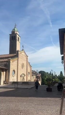 This is a vertical video taken from a low angle, showing a European town square on a sunny day with a clear blue sky and scattered white clouds. The video begins with a wide shot of a brick church with a bell tower and arched windows. Several cars are parked in front of the church, and a person is walking across the square. The camera then pans to the right, showing more of the church's facade and a covered walkway with arches.

The scene shifts to a view of the town square with more buildings, including a multi-story building with green shutters and balconies. Several cars are driving through the square, and a construction crane is visible in the background. The video then shows a white car turning into a street lined with buildings, one of which has a red awning and tables set up outside.

The camera continues to pan, revealing a building with the name "GILDA" above arched entrances. There are tables and umbrellas set up outside this building, suggesting it is a restaurant or cafe. A person in a blue shirt is walking away from the camera in front of this building. The video ends with a shot of the cobblestone square and the buildings surrounding it. The overall atmosphere is peaceful and leisurely.